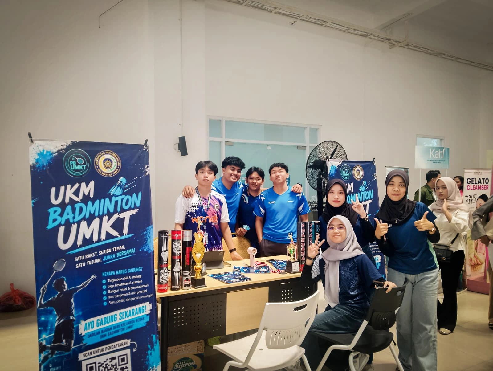 Pengurus UKM Badminton UMKT berpose di depan stan mereka, dengan spanduk bertuliskan Satu Raket Seribu Teman Satu Tujuan Juara Bersama, piala, dan tabung kok di atas meja