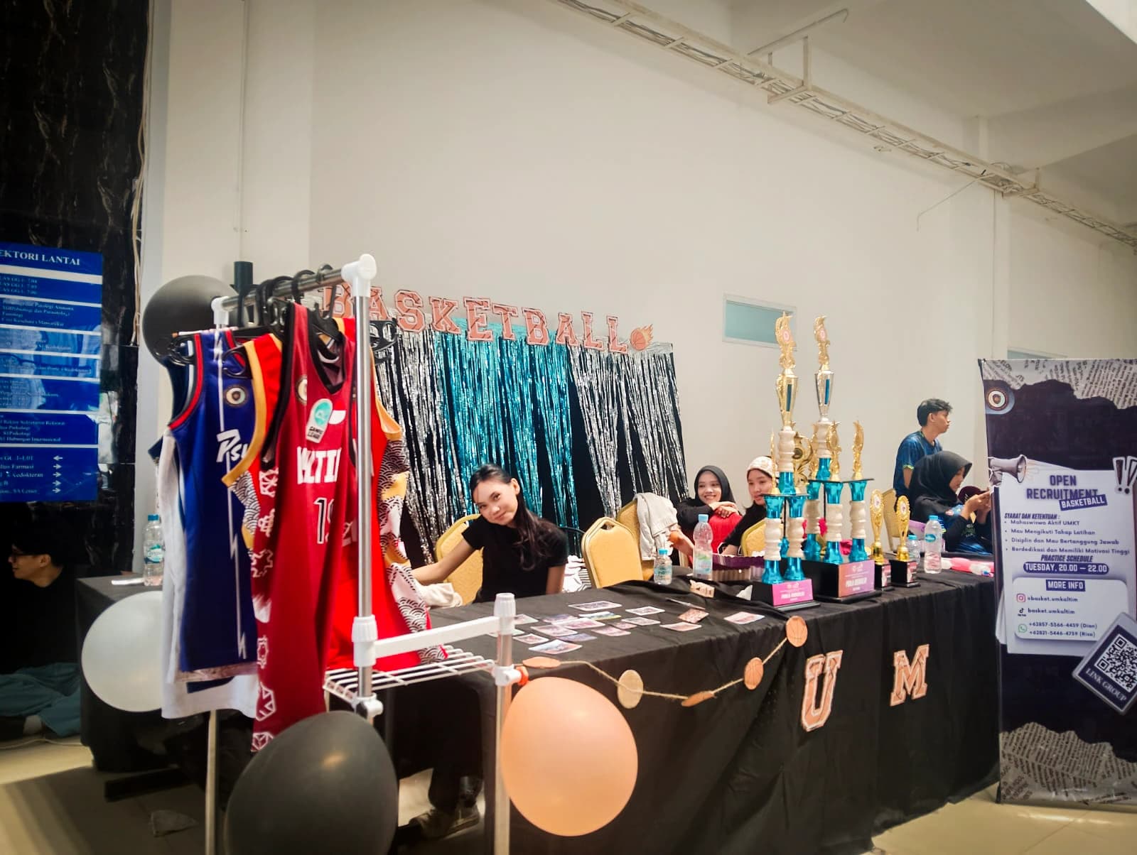 Stan UKM Basket dengan jersey tergantung di rak, deretan piala di atas meja berhias, dan spanduk bertuliskan Open Recruitment Basketball