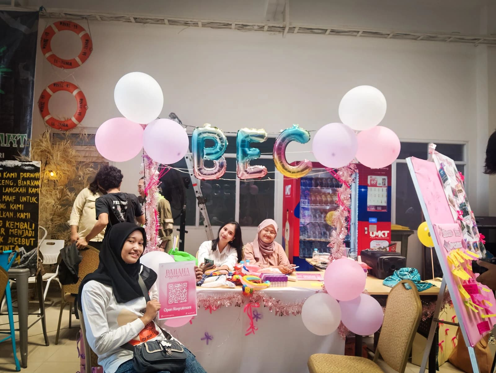 Stan paduan suara Bahana Enggang Choir berhias balon merah muda dan huruf balon BEC, dengan pengurus duduk di meja berselimut kain putih