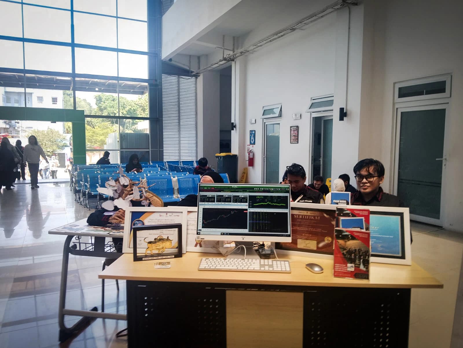 Stan kelompok studi pasar modal dengan monitor menampilkan grafik perdagangan saham, sertifikat berbingkai, dan patung banteng kecil di atas meja