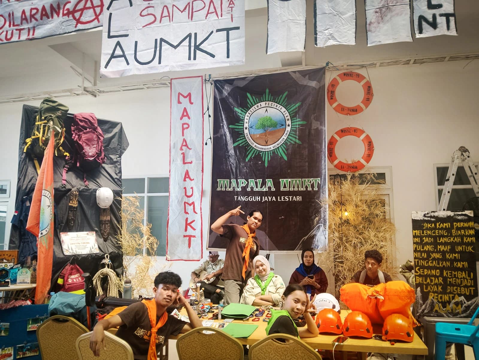 Stan MAPALA UMKT dengan spanduk bertuliskan Mapala UMKT Tangguh Jaya Lestari, dikelilingi tas gunung, helm panjat oranye, tali, dan pelampung
