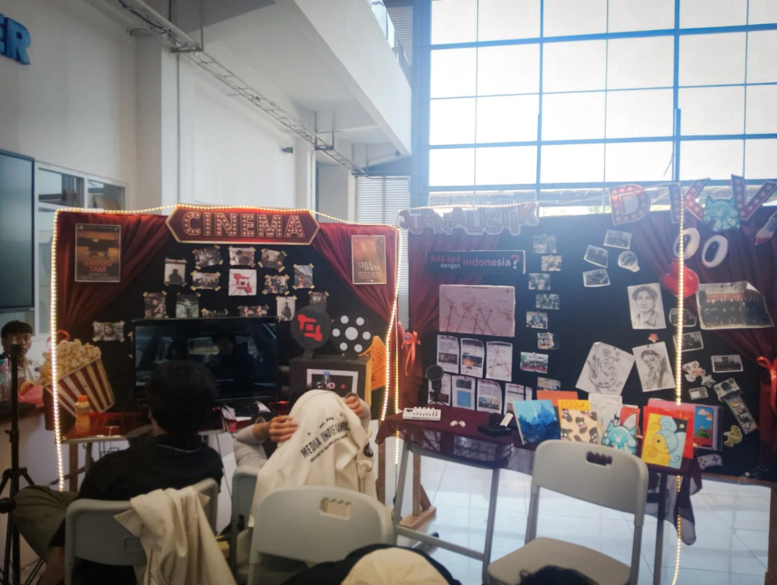 Stan Media Indie Kampus dengan dua panel bertema Cinema dan Jurnalistik, dihiasi lampu tumblr, poster film, dan papan berisi kliping serta karya ilustrasi