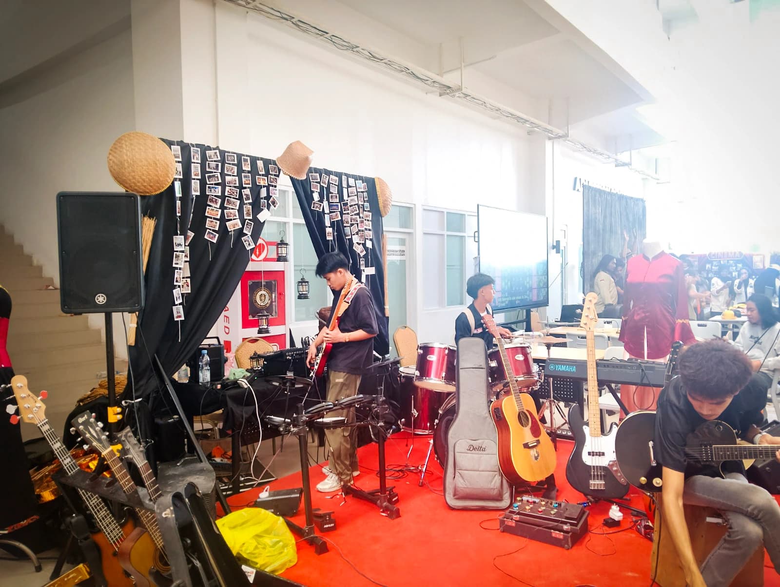 Stan unit seni dengan satu set alat musik lengkap di atas karpet merah: gitar, bas, drum, dan keyboard, berlatar kain hitam berhias foto polaroid dan caping anyaman