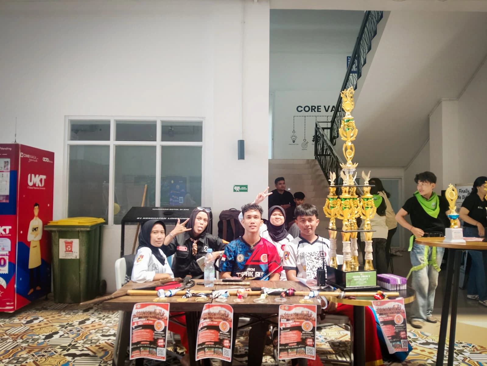 Pengurus UKM Tapak Suci berpose di belakang meja berisi tongkat dan senjata latihan, dengan piala bertingkat tinggi di sampingnya