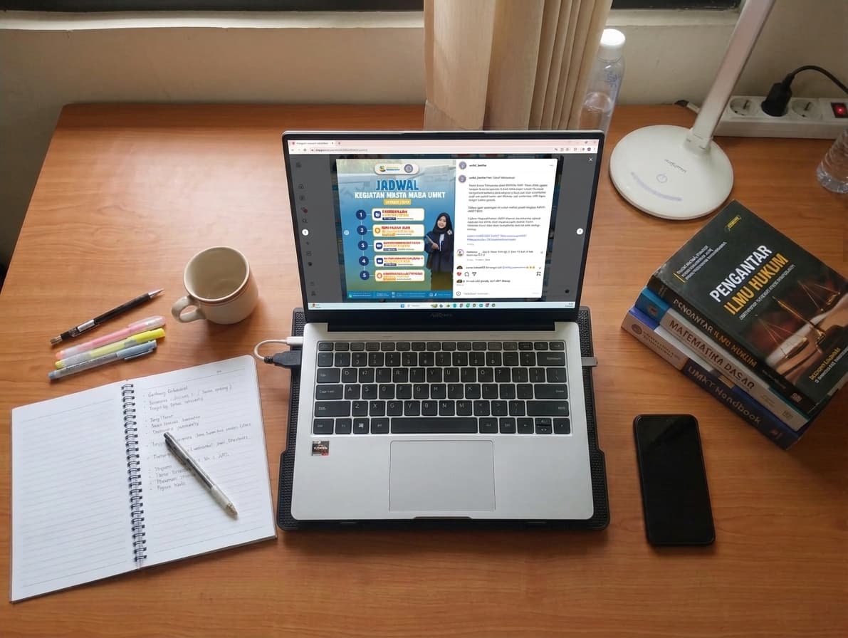Meja belajar dilihat dari atas: laptop menampilkan unggahan berisi poster jadwal kegiatan MASTA MABA UMKT dengan lima tahapan bernomor, di sampingnya buku catatan terbuka berisi tulisan tangan, pena, stabilo, cangkir, dan tumpukan buku kuliah