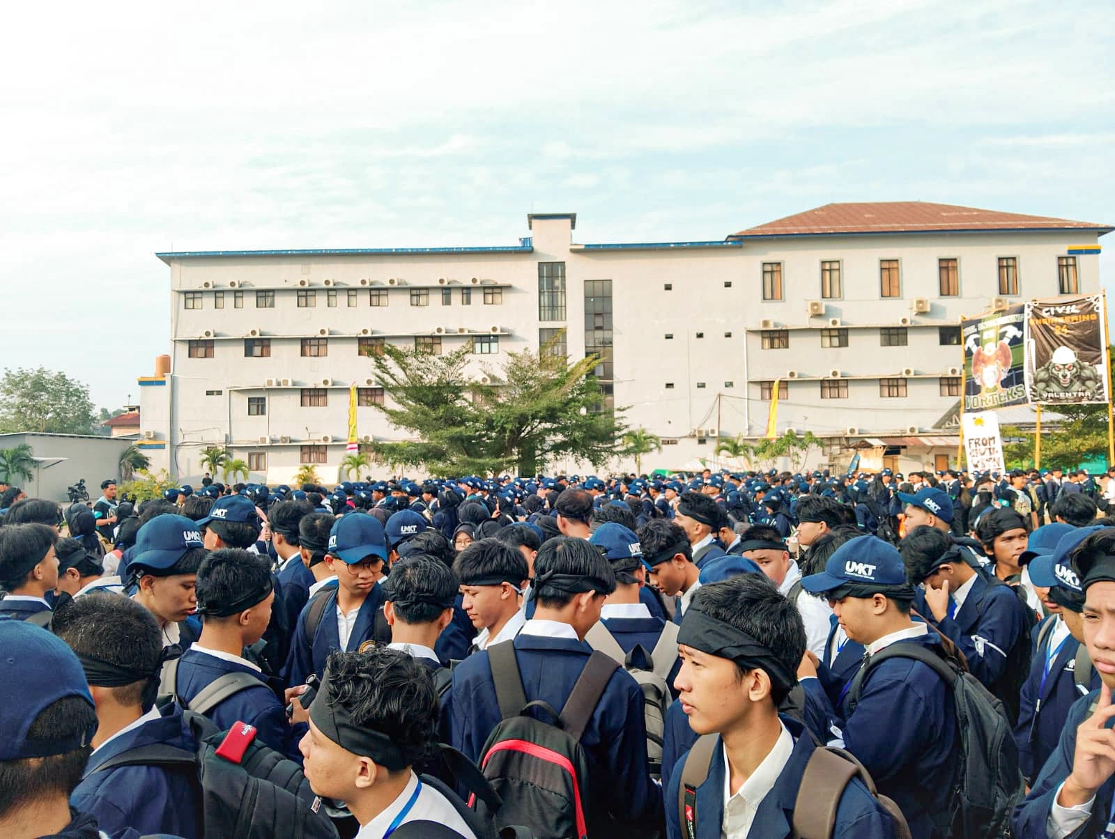Ribuan mahasiswa baru berjas almamater biru dan topi UMKT memadati lapangan kampus, dengan gedung perkuliahan dan spanduk program studi di latar belakang