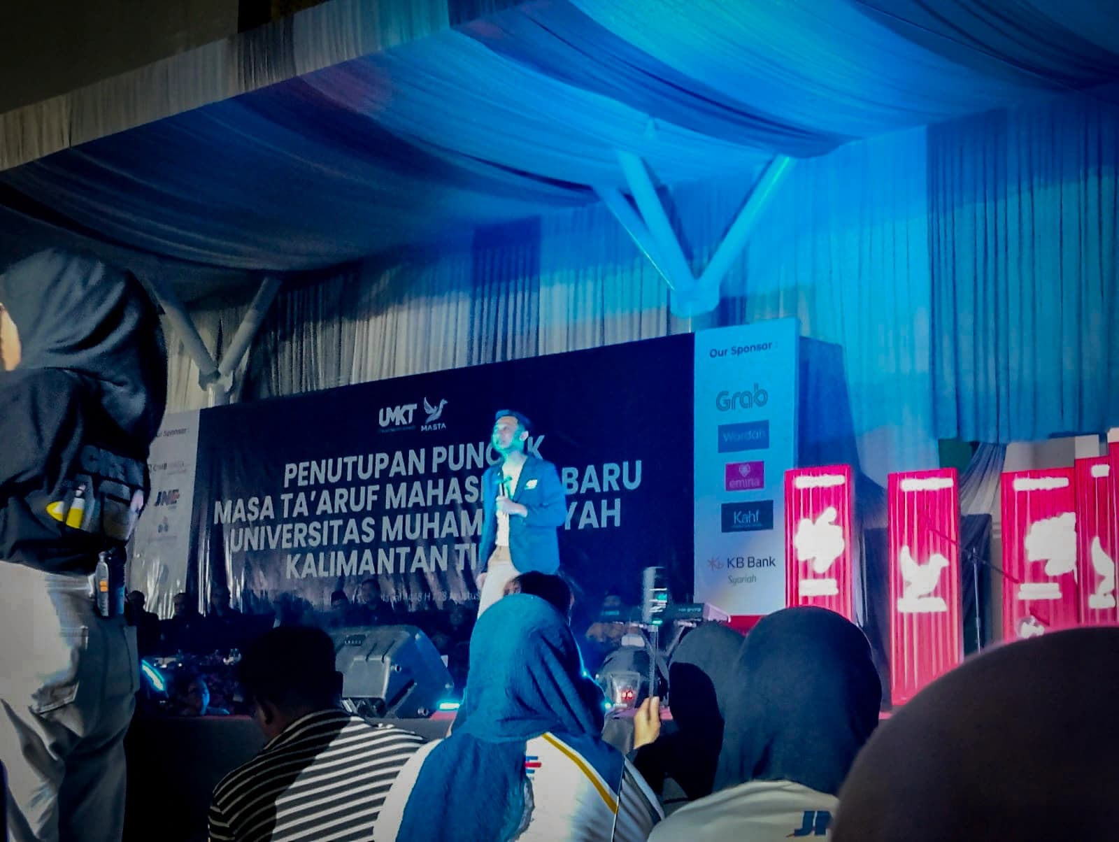 Bintang tamu berjas biru memegang mikrofon di panggung malam puncak di bawah lampu sorot biru, dengan penonton menyiluet di depan