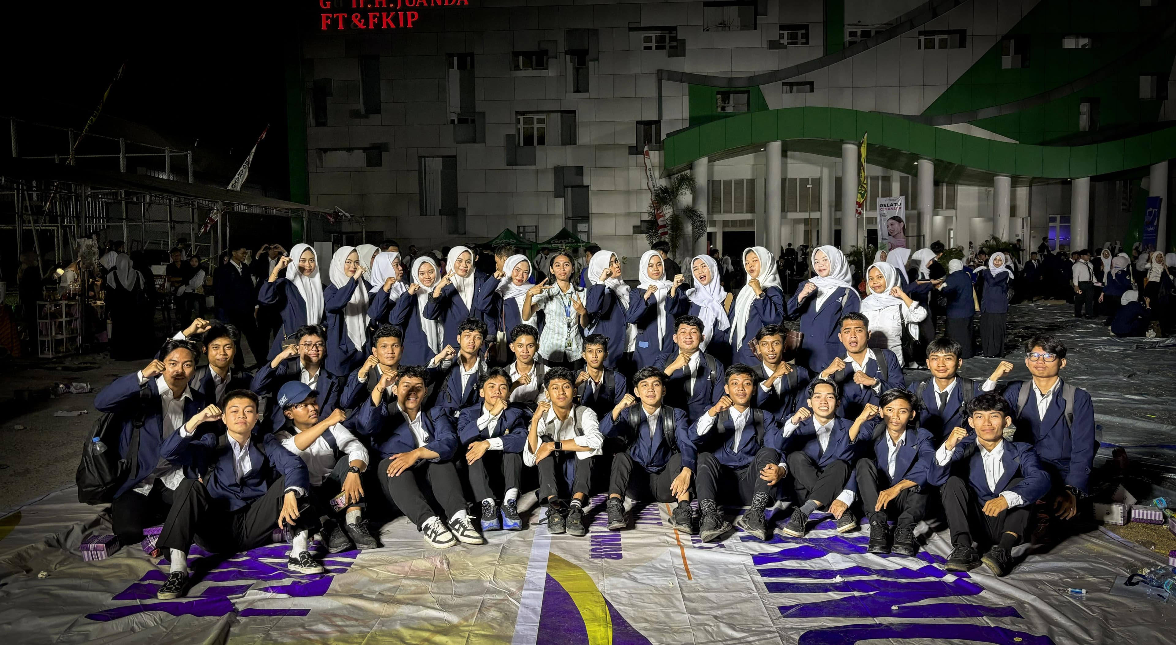 Sekitar tiga puluh lima mahasiswa baru berjas almamater biru berfoto bersama sambil mengepalkan tangan pada malam hari, di depan gedung bertuliskan Gedung H. Juanda FT dan FKIP