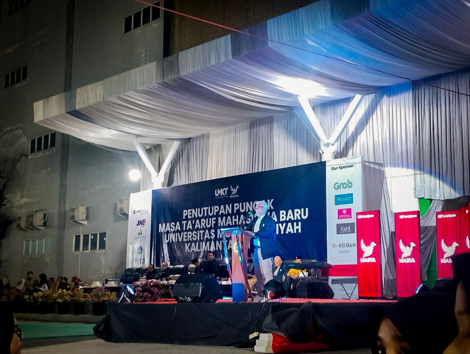 Rektor berdiri di podium panggung malam puncak menyampaikan pesan penutup, dengan spanduk penutupan MASTA dan jajaran tamu undangan di belakangnya