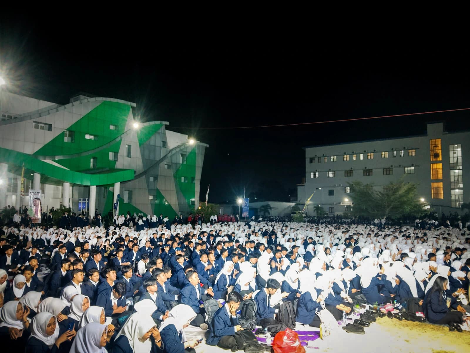 Ribuan mahasiswa baru berjas almamater biru duduk lesehan memenuhi lapangan kampus pada malam hari, dengan gedung perkuliahan berlampu di belakang mereka