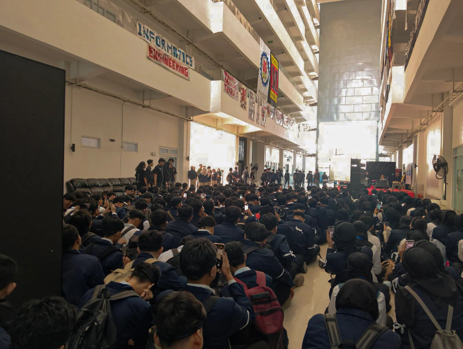 Ratusan mahasiswa baru duduk lesehan memenuhi selasar gedung fakultas sambil merekam dengan ponsel, spanduk himpunan program studi tergantung di lantai atas