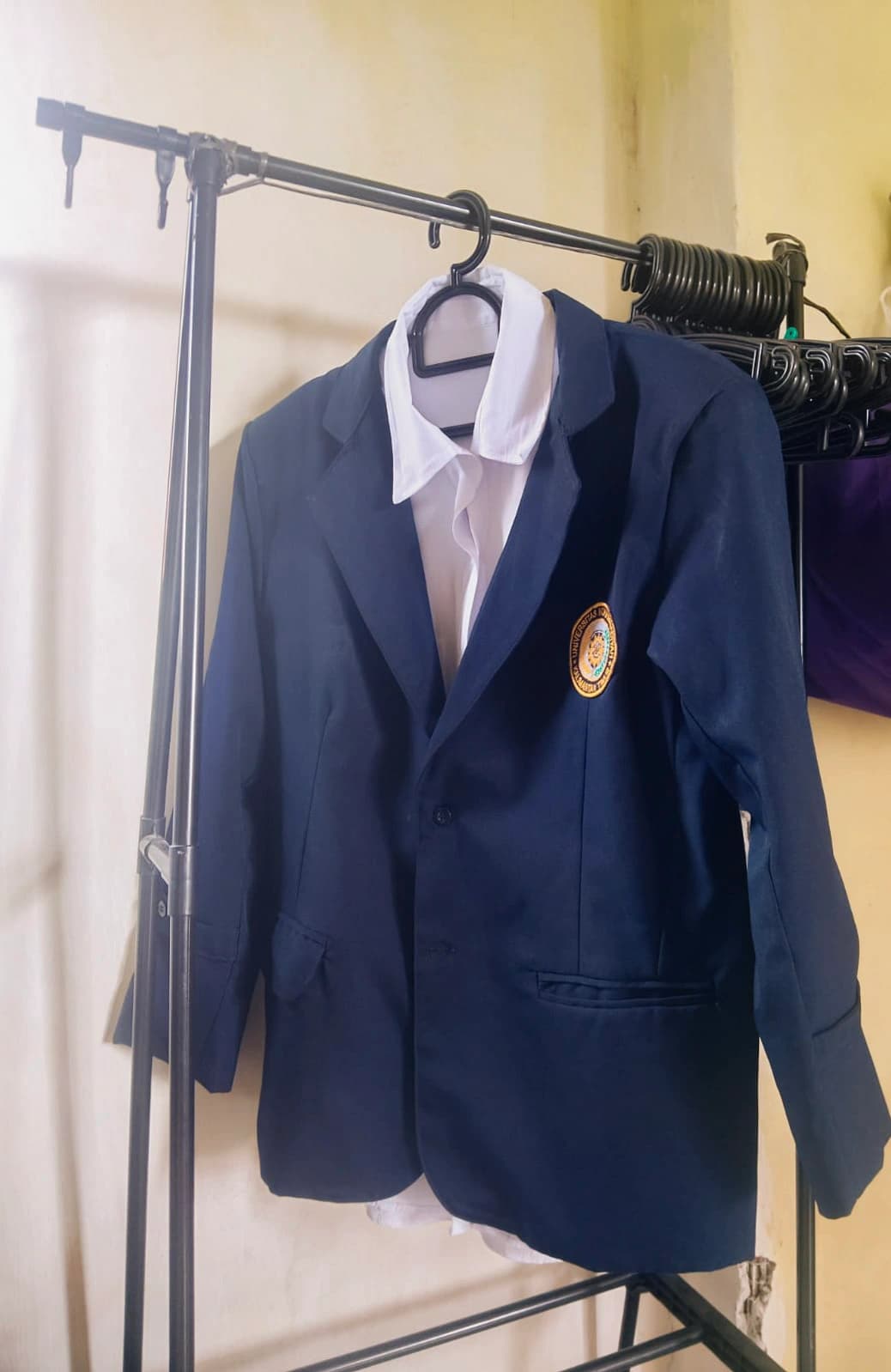 Jas almamater biru tua bersulam lambang UMKT tergantung di rak baju bersama kemeja putih, dengan dinding polos sebagai latar