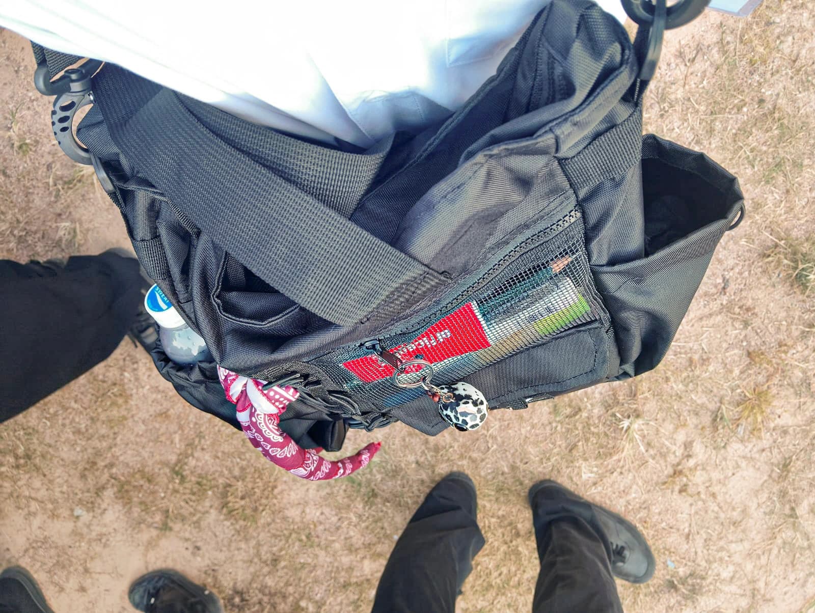 Pandangan dari atas ke tas selempang hitam yang sedang dipakai, dengan botol minum di kantong samping dan bandana merah terikat di talinya, berlatar rumput kering