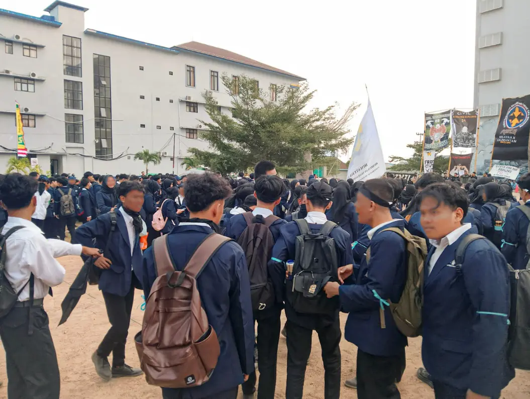 Kerumunan mahasiswa baru berjas almamater biru berdiri di lapangan kampus, sebagian saling mengobrol, dengan bendera dan spanduk himpunan program studi berkibar di latar belakang