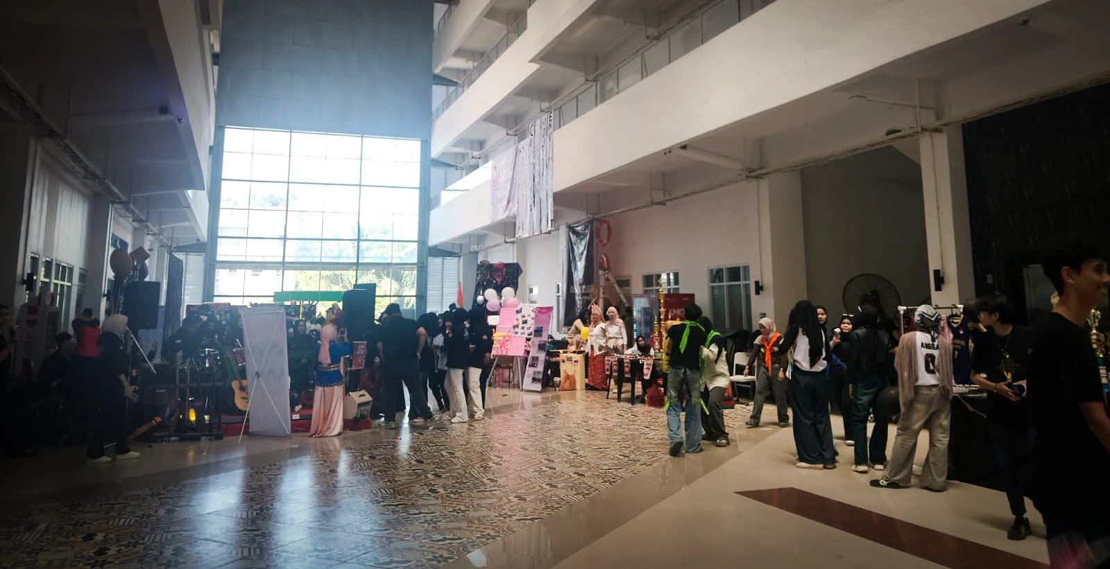 Selasar gedung kampus yang dipenuhi deretan stan unit kegiatan mahasiswa saat UKM Expo, dengan mahasiswa berjalan dan berkerumun di antara stan