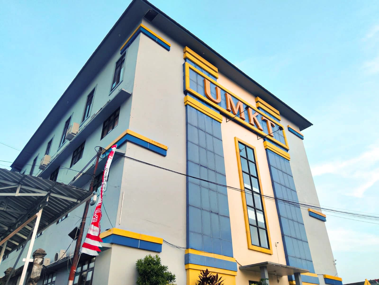 Gedung kampus UMKT difoto dari sudut rendah, fasad biru-kuning dengan huruf timbul UMKT berwarna emas di bawah langit cerah