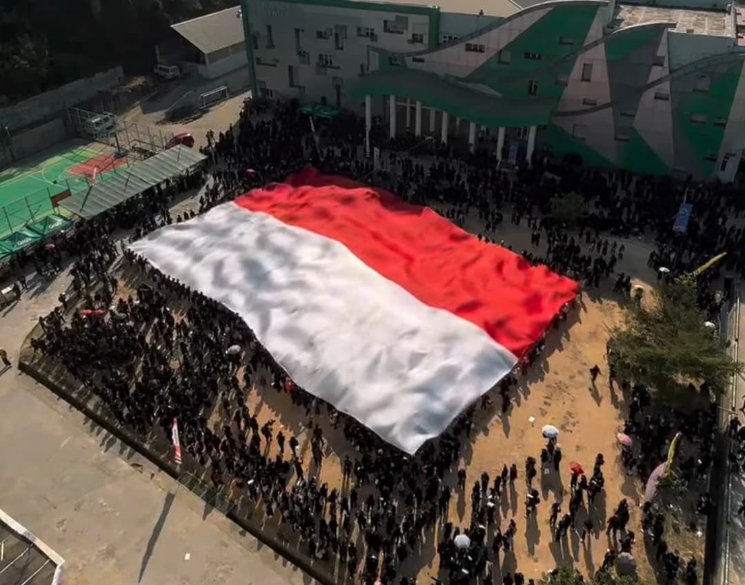 Bendera Merah Putih berukuran raksasa dibentangkan mendatar di lapangan kampus, dikelilingi ratusan mahasiswa yang memegang tepinya, difoto dari atas